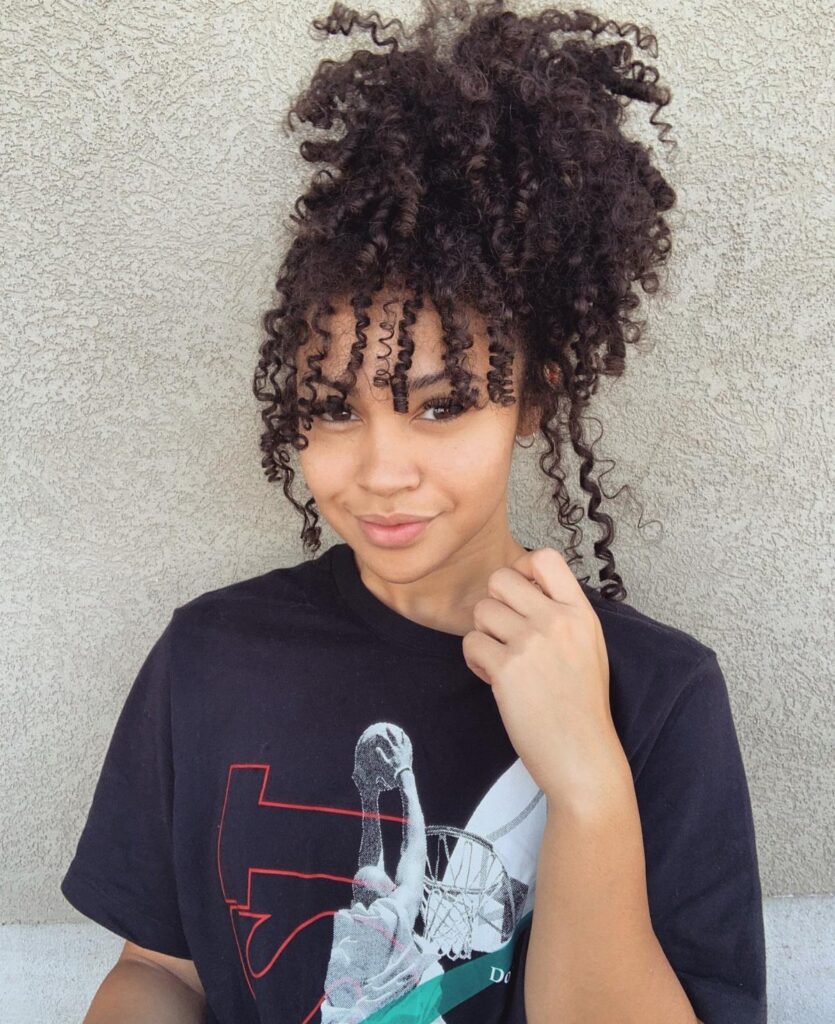 Woman with dark curls gathered high in a pineapple ponytail, loose ringlets framing her face