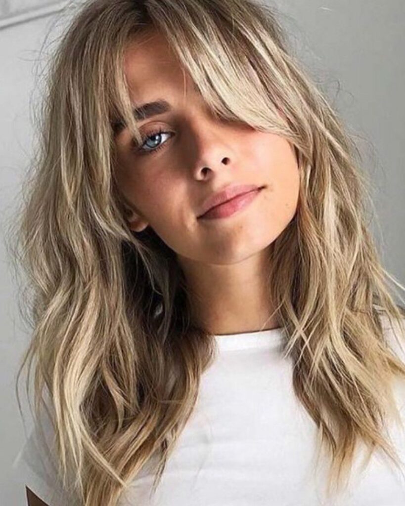 Woman with long layered blonde waves and sweeping bangs against a light wall