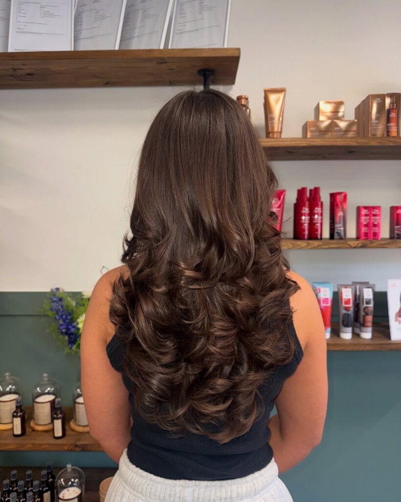 Long dark brown layered hair styled in loose curls, seen from behind in a salon