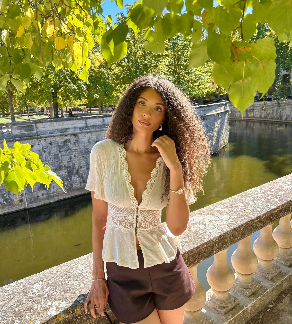 Woman with long dark curls poses in a white lace blouse by a canal