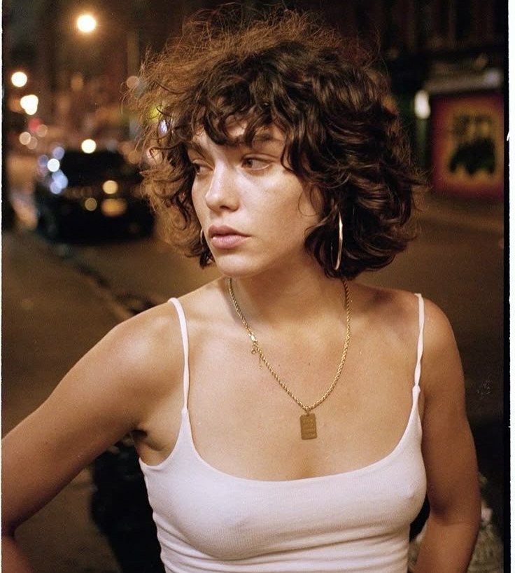 Woman with a short curly bob and bangs poses on a city street at night.