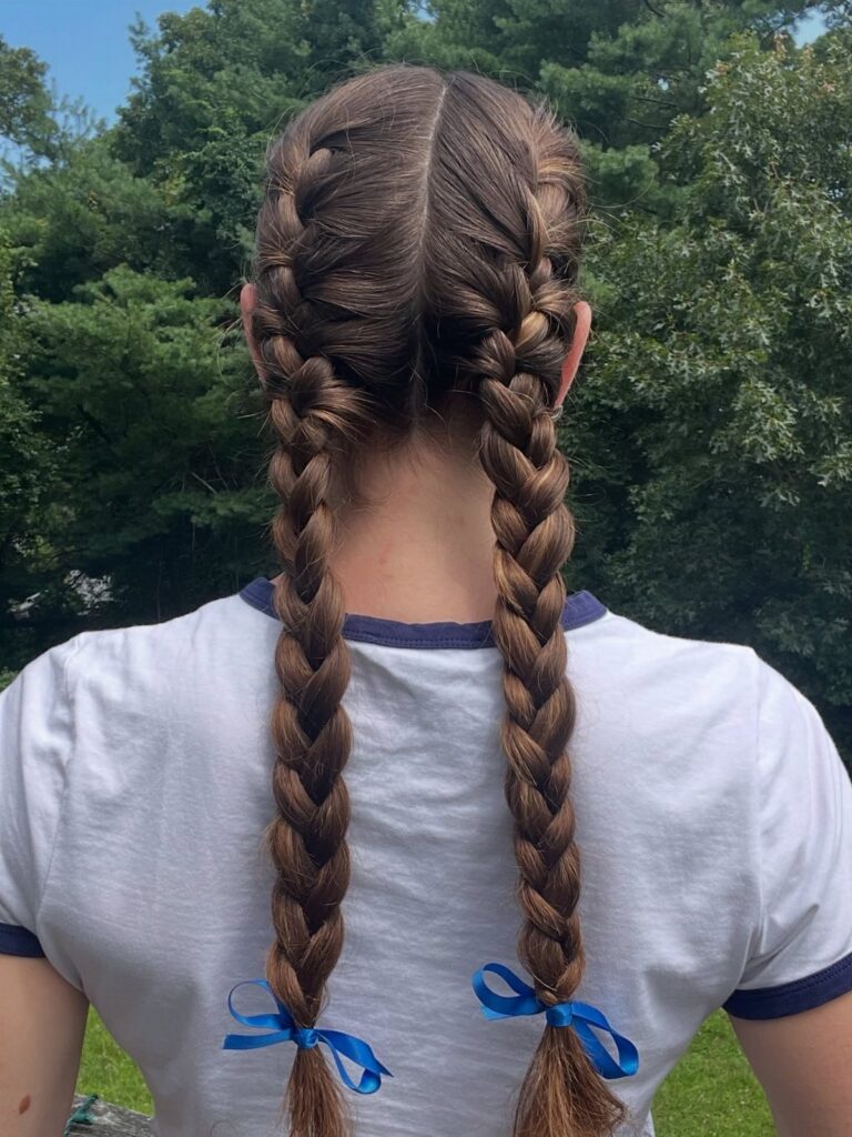 Two long French braid pigtails tied with blue ribbons against a leafy outdoor backdrop